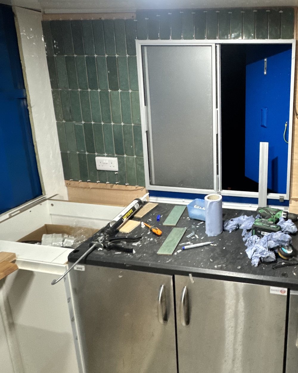 Green tiles going up around a sliding hatch above stainless fridges