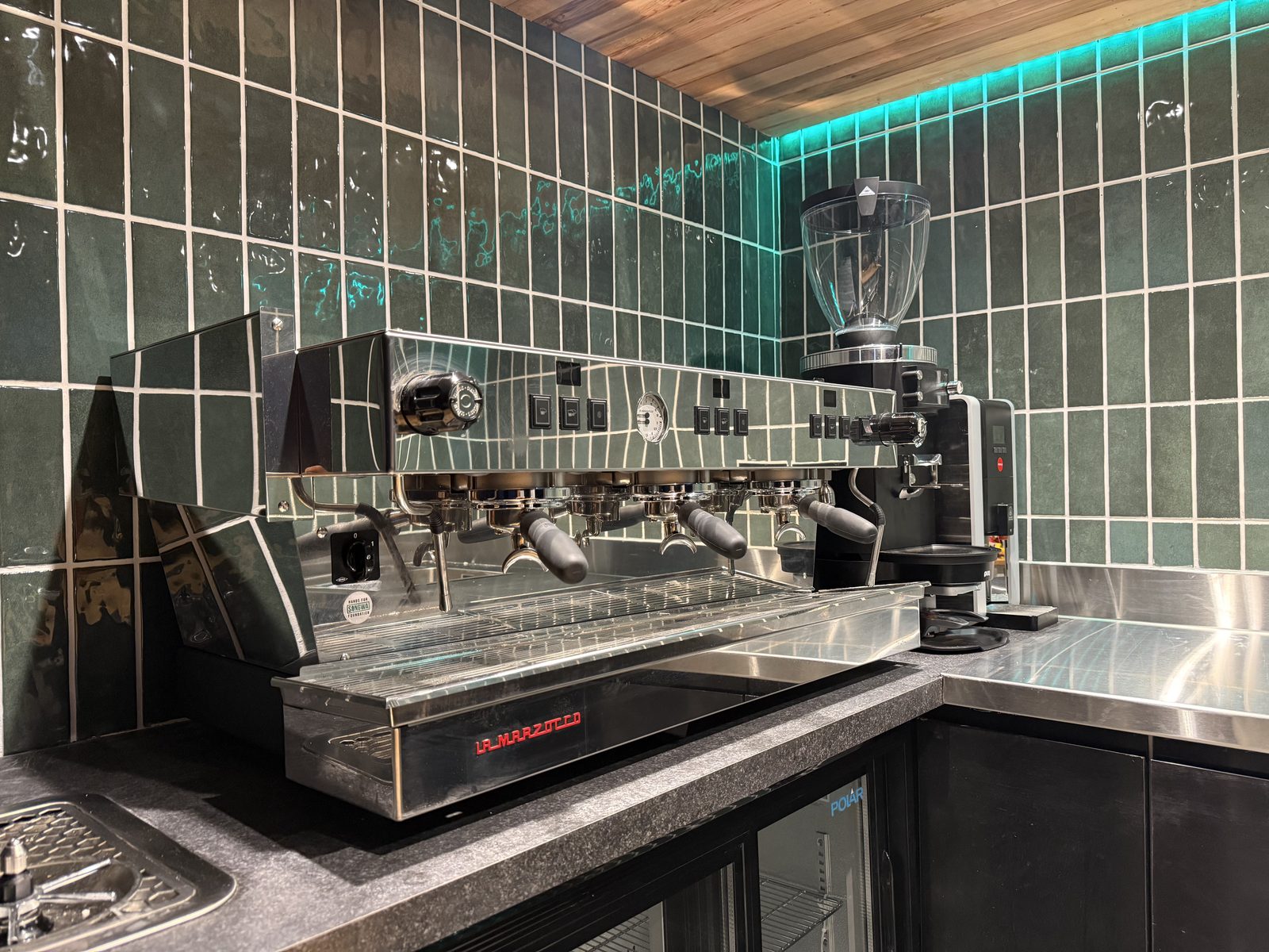 A commercial espresso machine and grinder on a stainless counter against green tiles