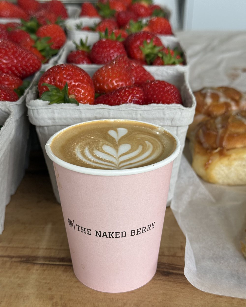 A latte in a pink Naked Berry cup in front of punnets of fresh strawberries