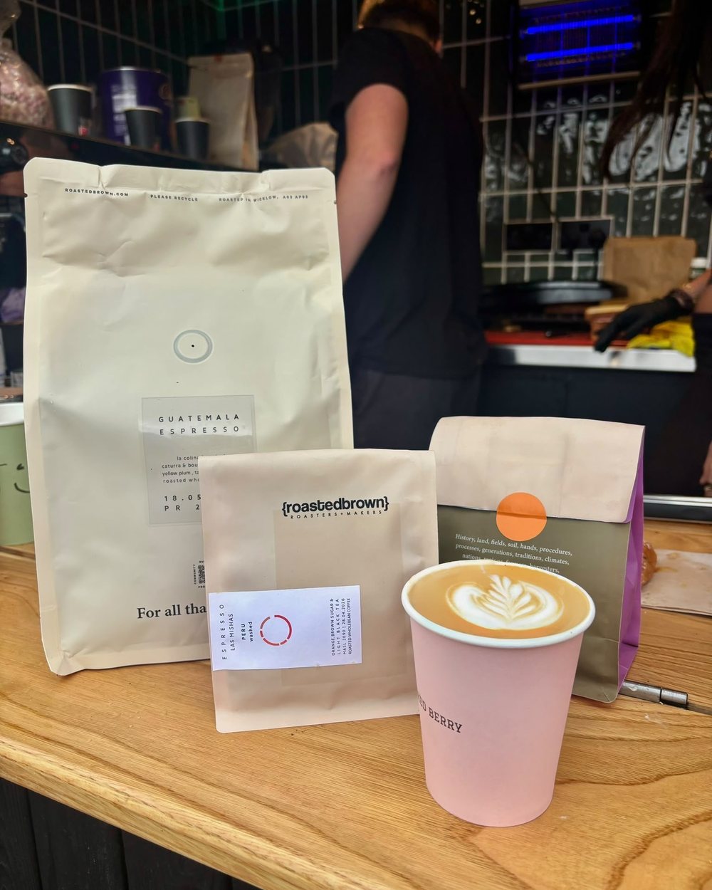 A latte with rosetta art beside bags of Roasted Brown espresso beans